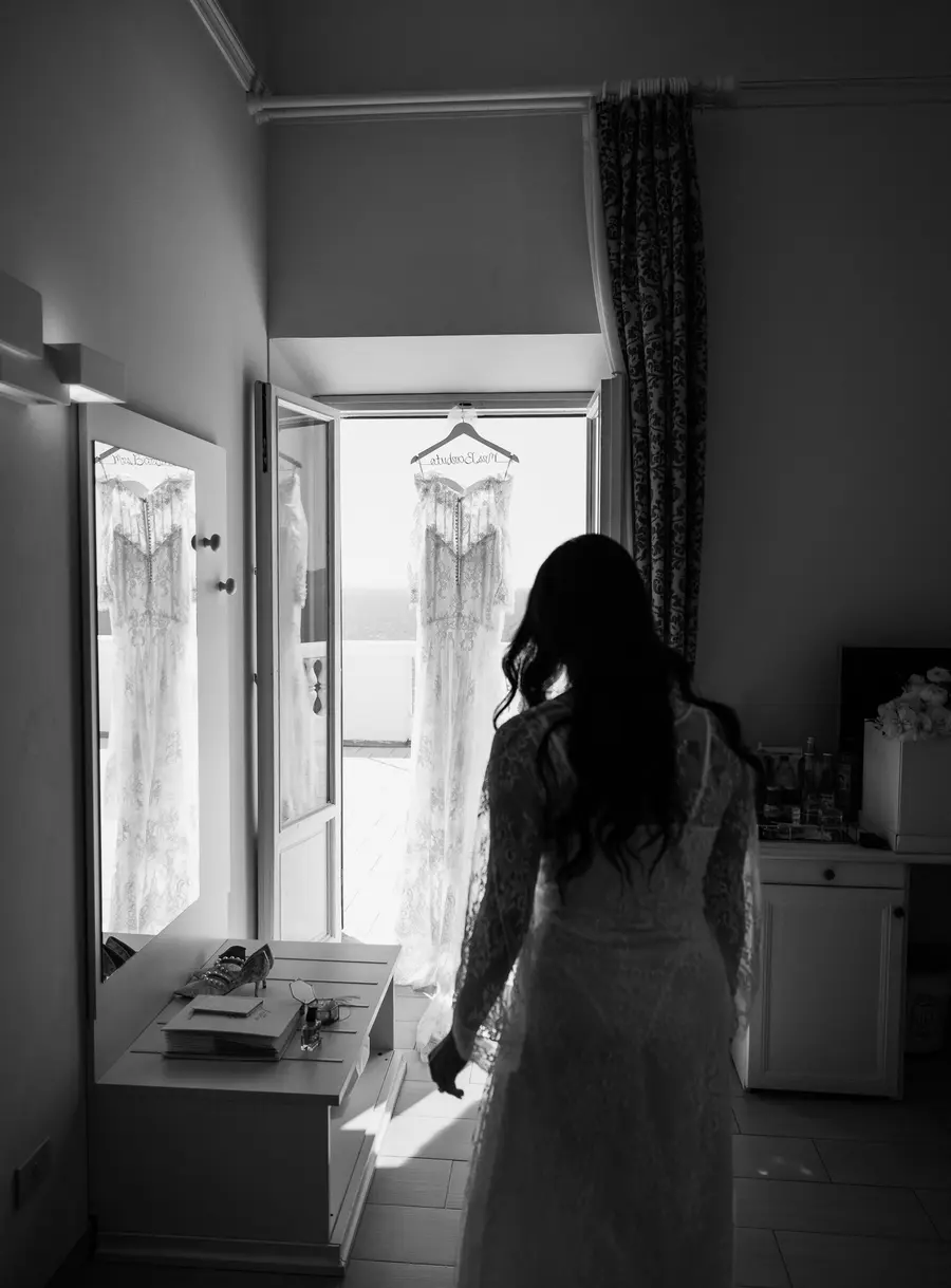 La sposa in vestaglia si avvicina all'abito da sposa appeso alla porta finestra, bianco e nero
