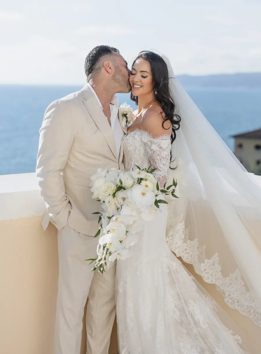 Bacio sulla guancia della sposa con bouquet di orchidee e velo di pizzo sul mare