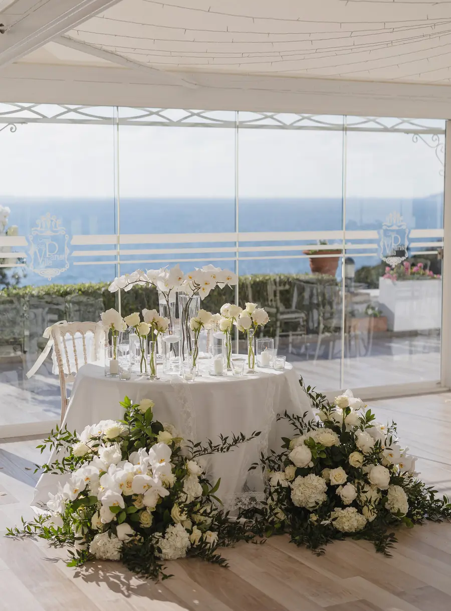 Tavolo degli sposi con vasetti di orchidee e fiori bianchi nel padiglione vetrato sul mare