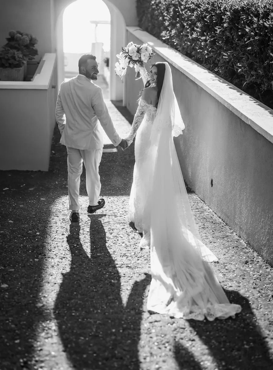 Gli sposi mano nella mano nel vialetto in controluce, la sposa si volta, bianco e nero