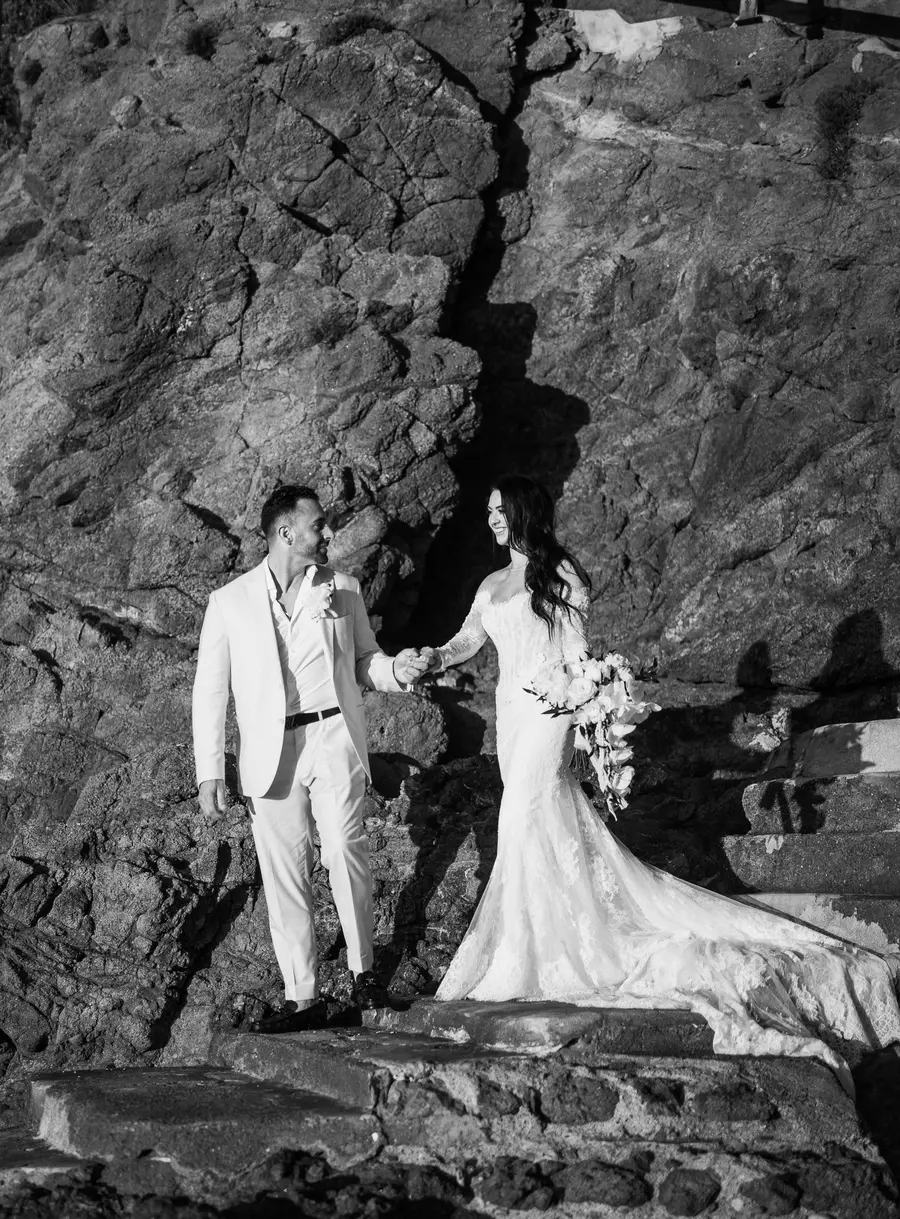 Gli sposi si tengono per mano tra le rocce con uno sguardo complice, bianco e nero