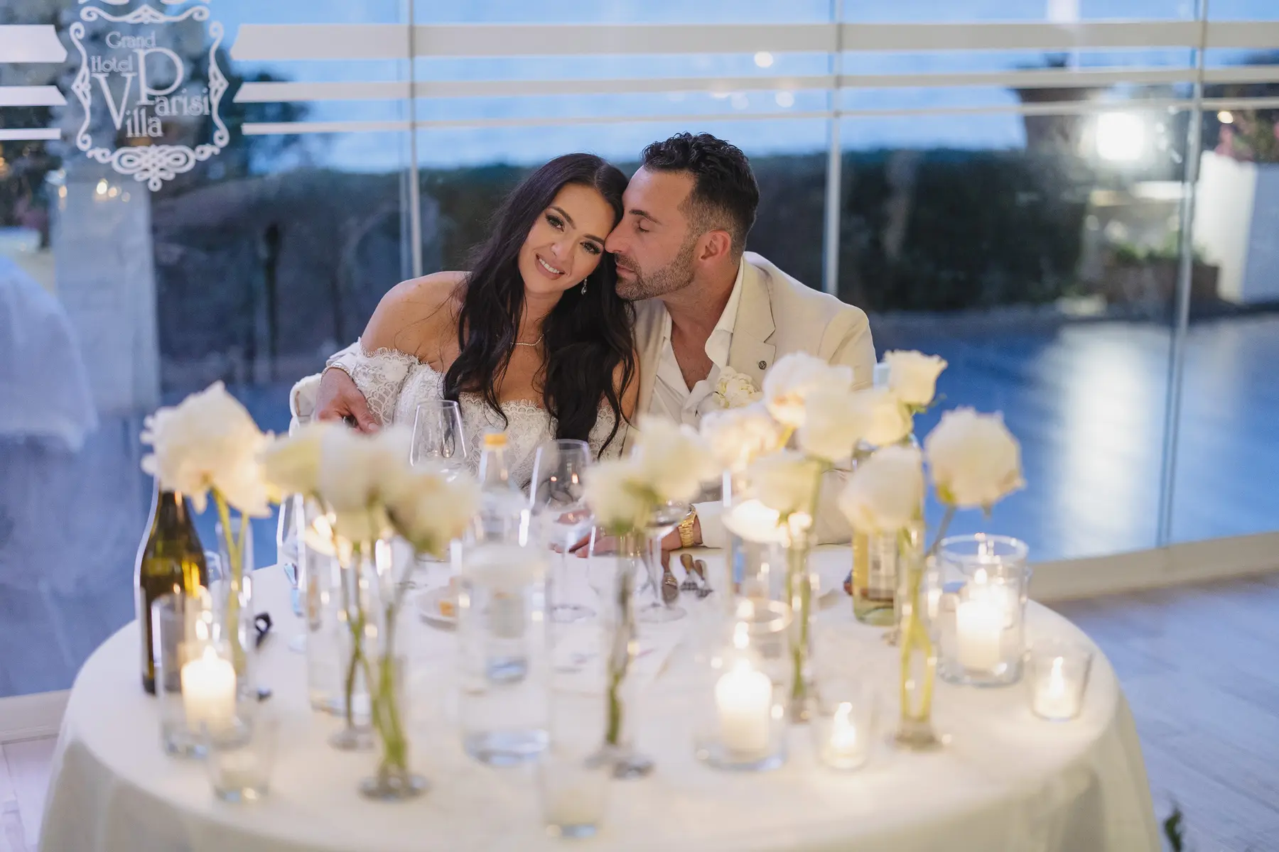 Gli sposi abbracciati al tavolo con candele e rose bianche a Villa Parisi