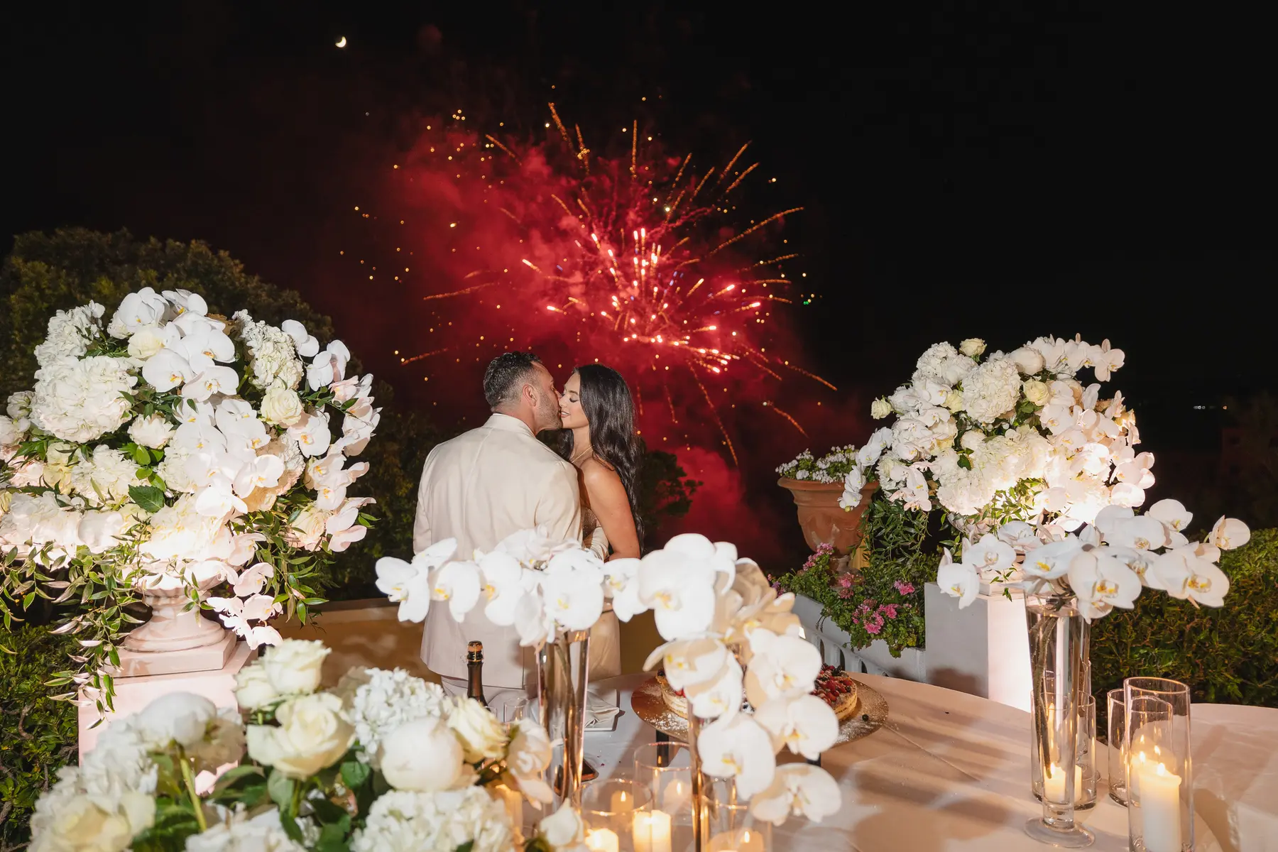 Gli sposi si baciano di notte tra fiori bianchi mentre i fuochi d'artificio illuminano il cielo